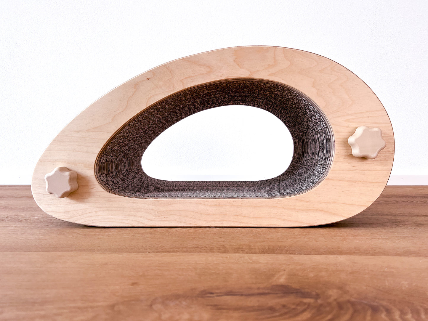 Kratzmöbel für Katzen in Form einer abstrakten Maus mit einem Liegetunnel im Inneren bestehend aus zwei hellen Holzelementen mit Wellpappebögen dazwischen. Die Verschraubung erfolgt mit Muttern aus Holzfilament in Form von abgerundeten Sternen.