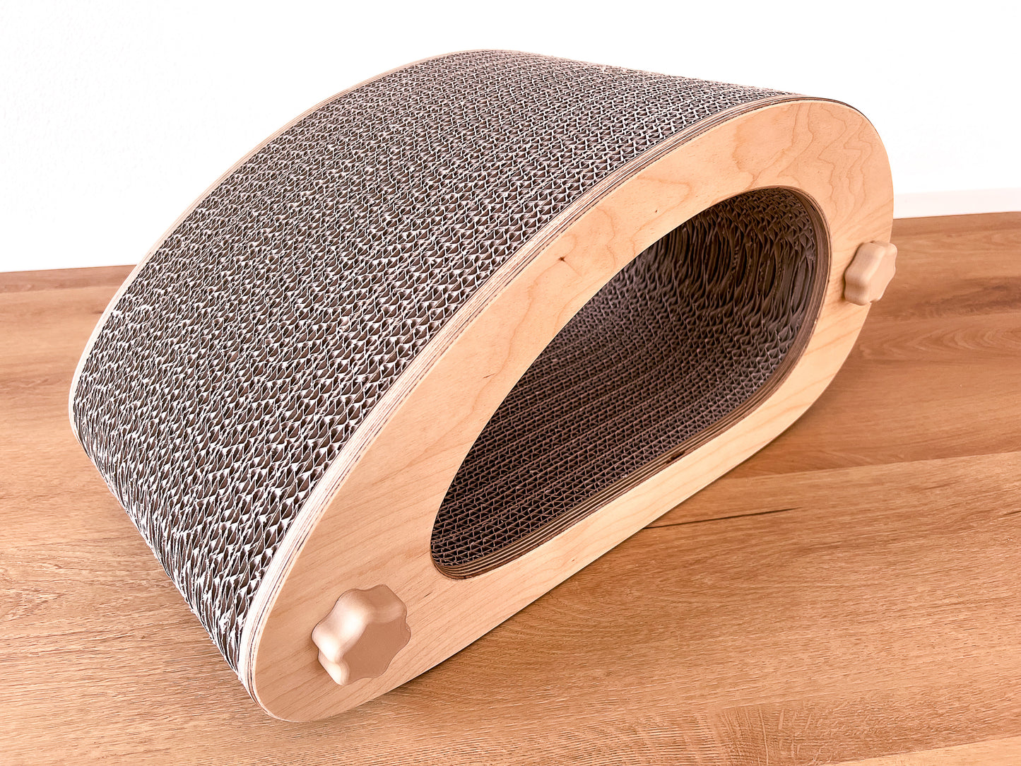 Kratzmöbel für Katzen in Form einer abstrakten Maus mit einem Liegetunnel im Inneren bestehend aus zwei hellen Holzelementen mit Wellpappebögen dazwischen. Die Verschraubung erfolgt mit Muttern aus Holzfilament in Form von abgerundeten Sternen.