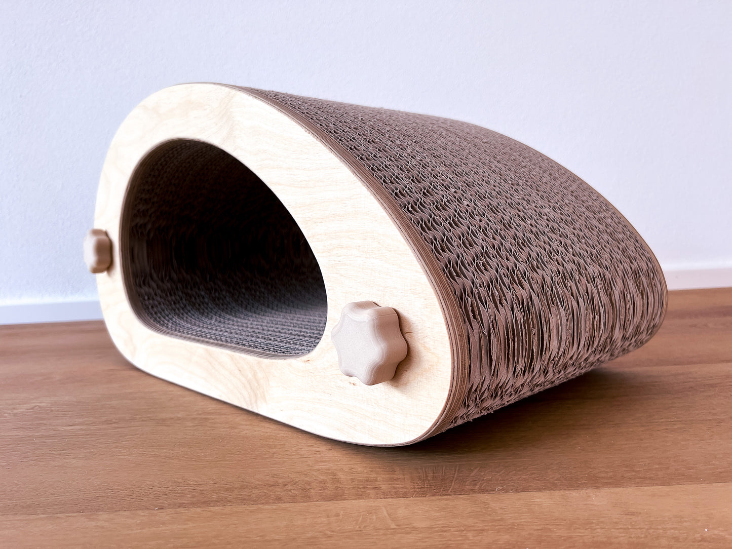 Kratzmöbel für Katzen in Form einer abstrakten Maus mit einem Liegetunnel im Inneren bestehend aus zwei hellen Holzelementen mit Wellpappebögen dazwischen. Die Verschraubung erfolgt mit Muttern aus Holzfilament in Form von abgerundeten Sternen.