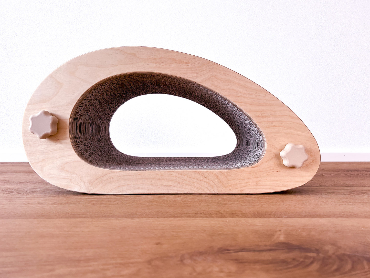 Kratzmöbel für Katzen in Form einer abstrakten Maus mit einem Liegetunnel im Inneren bestehend aus zwei hellen Holzelementen mit Wellpappebögen dazwischen. Die Verschraubung erfolgt mit Muttern aus Holzfilament in Form von abgerundeten Sternen.