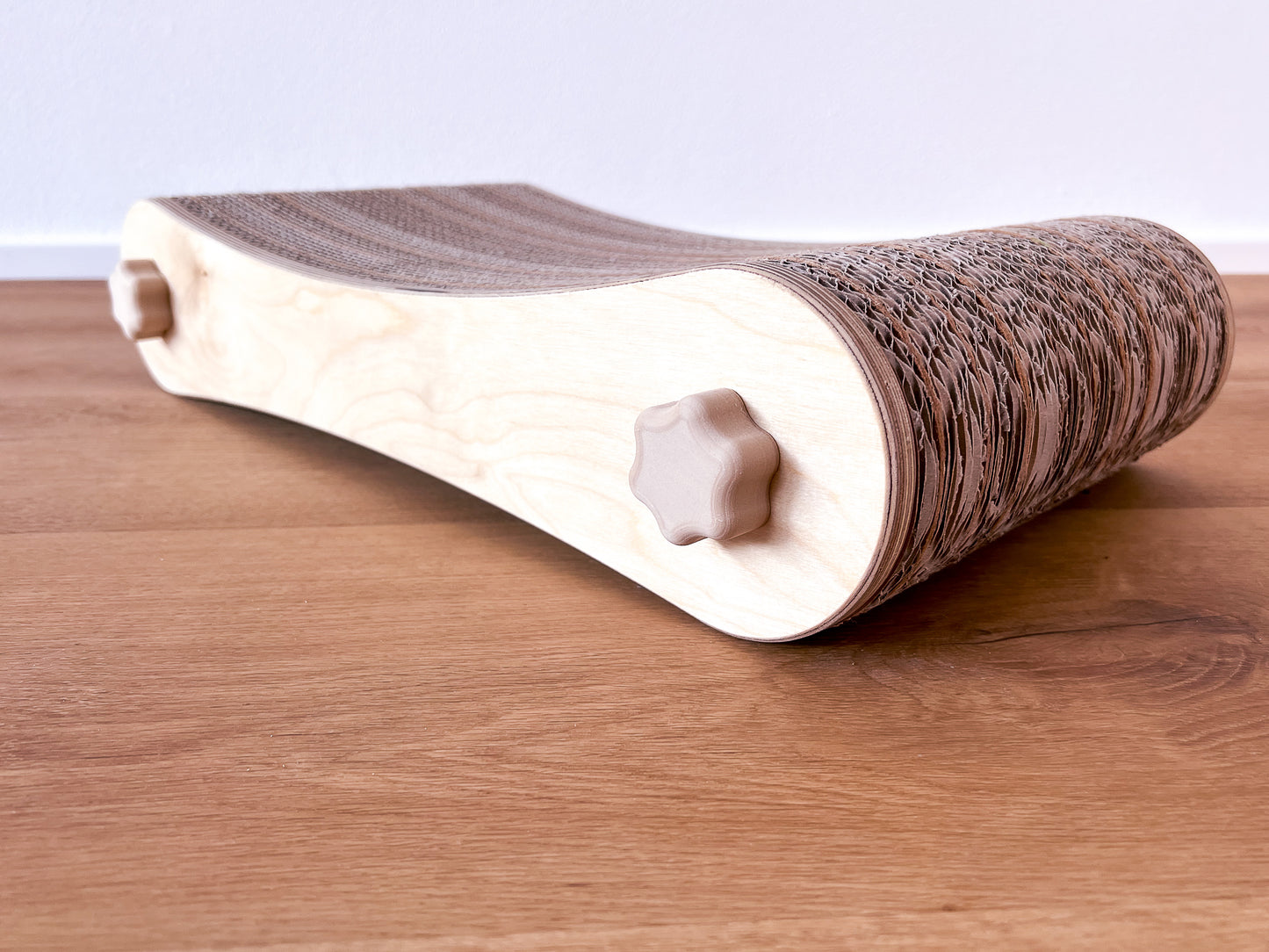Kratzmöbel für Katzen in Form einer Liege bestehend aus zwei hellen Holzelementen mit Wellpappebögen dazwischen. Die Verschraubung erfolgt mit Muttern aus Holzfilament in Form von abgerundeten Sternen.