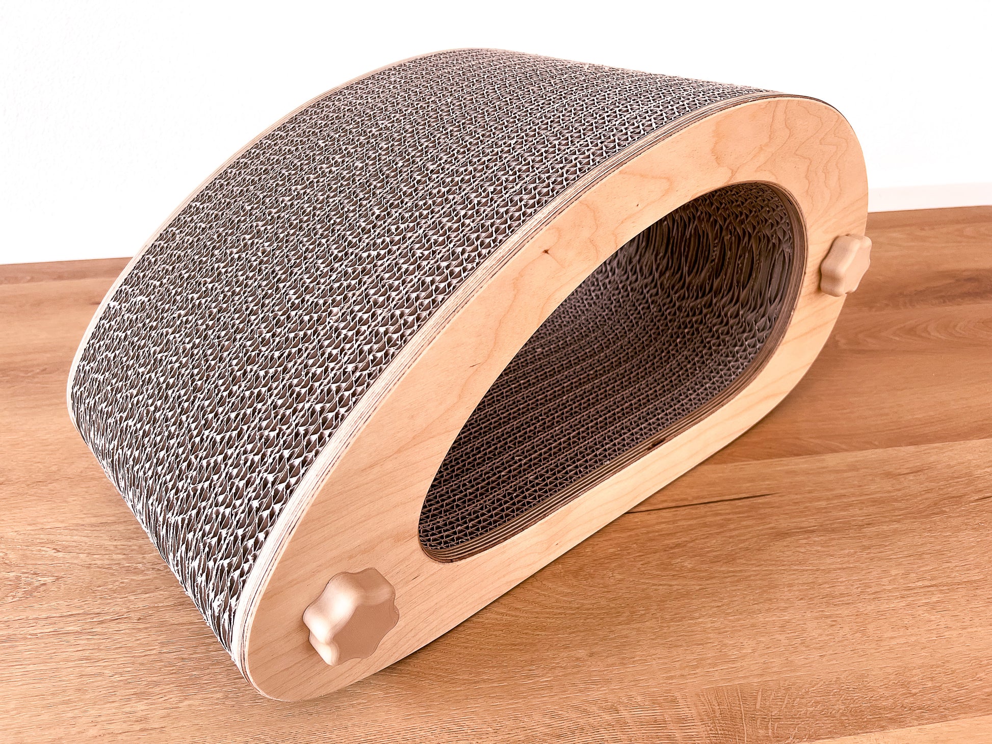 Kratzmöbel für Katzen in Form einer abstrakten Maus mit einem Liegetunnel im Inneren bestehend aus zwei hellen Holzelementen mit Wellpappebögen dazwischen. Die Verschraubung erfolgt mit Muttern aus Holzfilament in Form von abgerundeten Sternen.